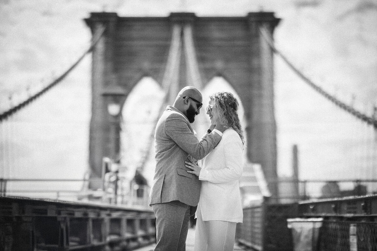 Black and white image of a couple standing close together on the Brooklyn Bridge after their NYC elopement, framed by the bridge’s arches and cables, highlighting the blend of iconic architecture and personal connection.