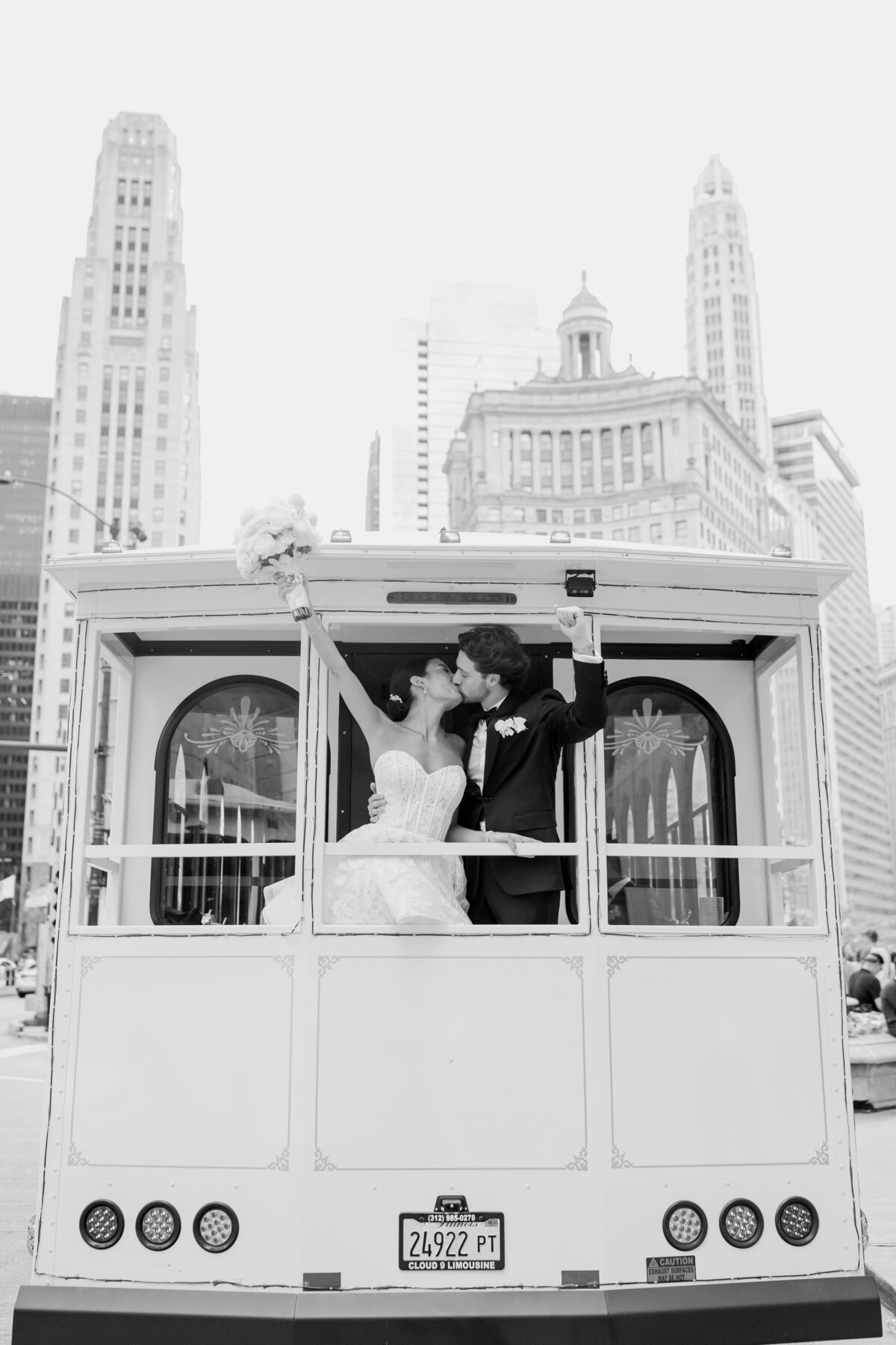 Bride and groom kiss on the back of a trolley car driving down a Chicago street after their wedding