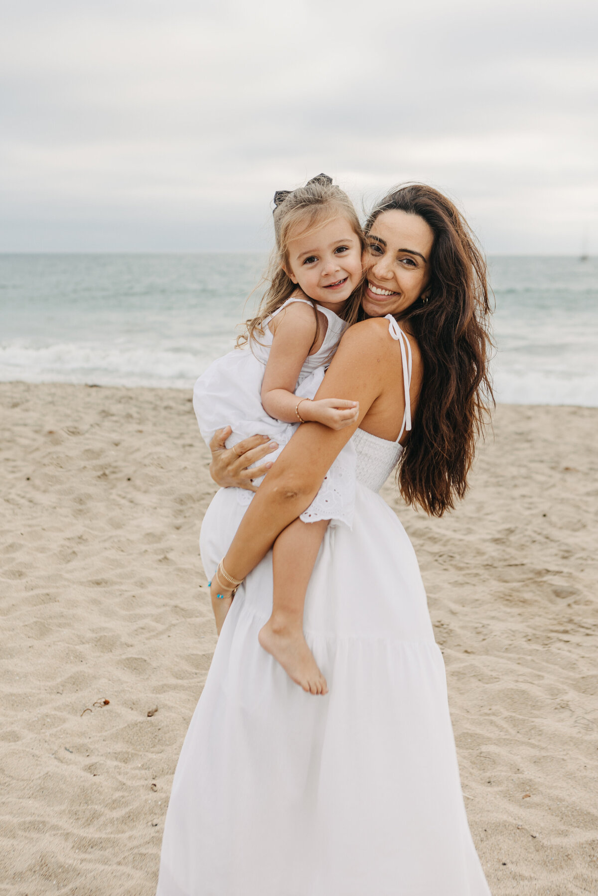 Mom in white dress holds her daughter while pregnant on the beach in Los Angeles