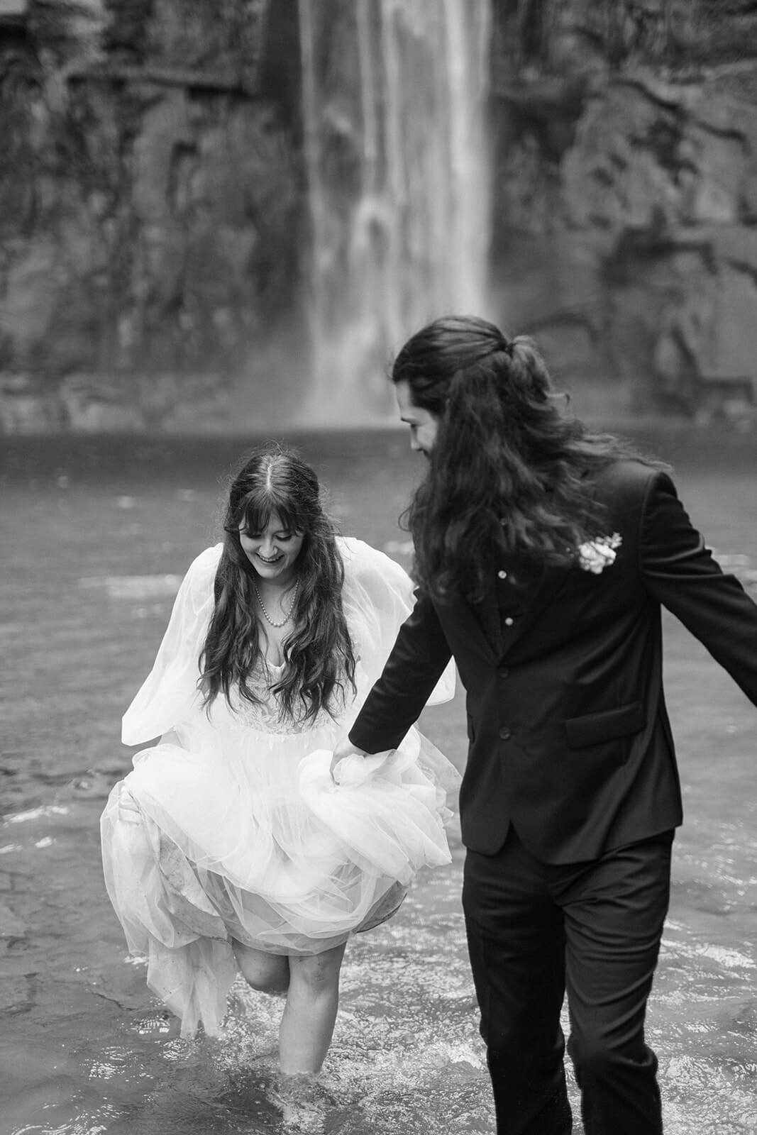 bride and groom walking through water in front of a waterfall