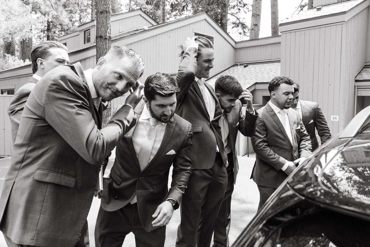 Seven groomsmen fix their hair in the reflection of a car window at their friend's wedding.