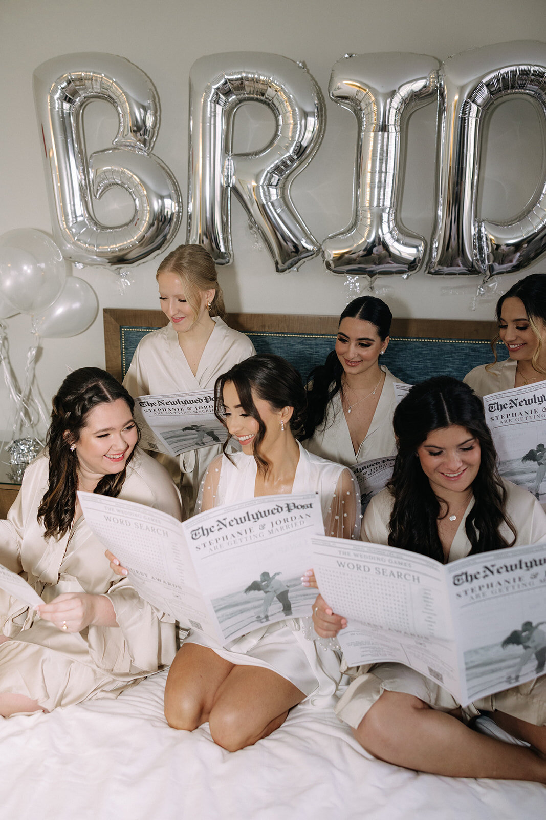 bride and bridesmaids sitting on bed reading newspaper announcements at bella collina wedding venue​