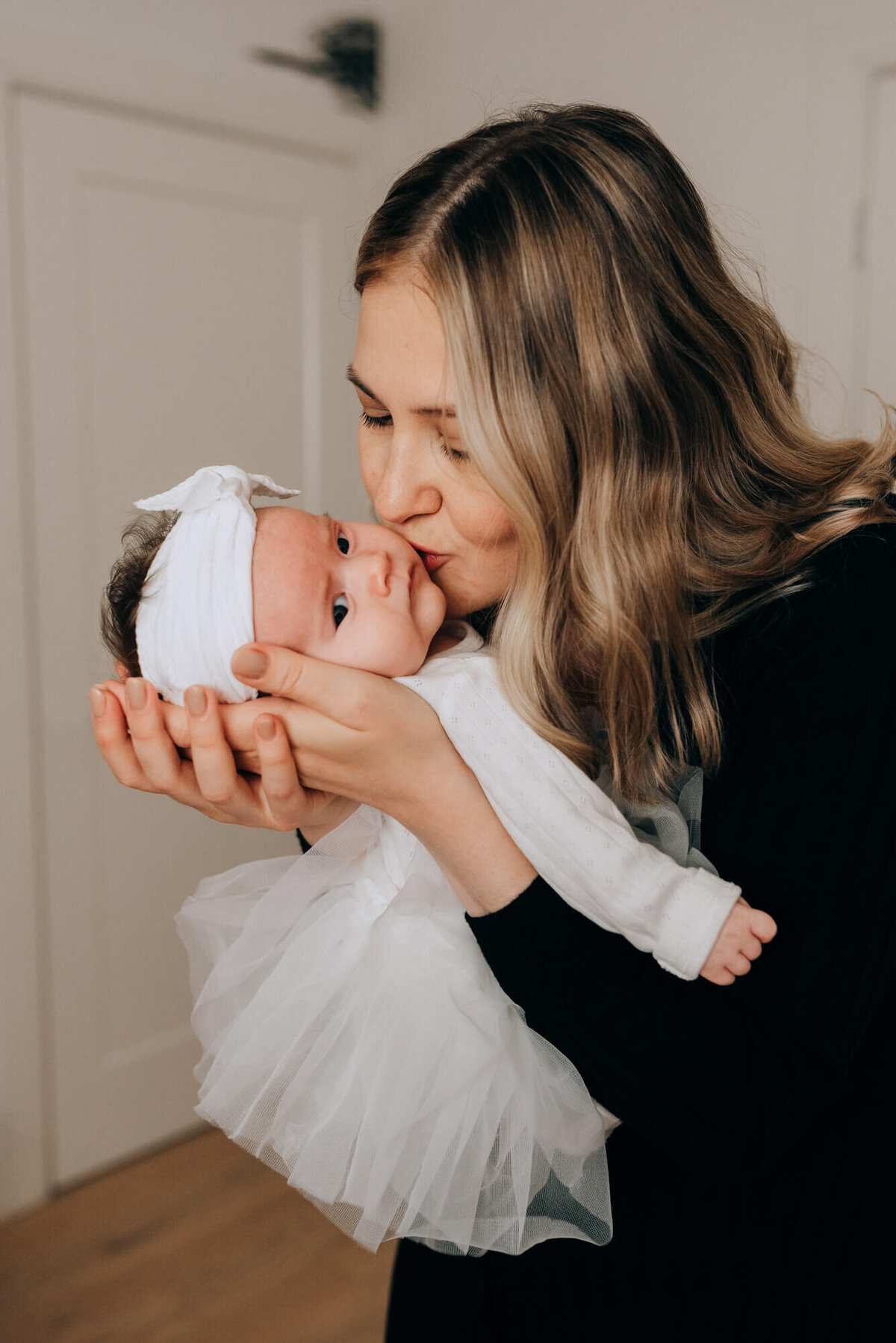 A mother wearing a sleek black dress kisses her newborn daughter on the cheek during their photoshoot with Allison Wolf, Boston Newborn Photographer