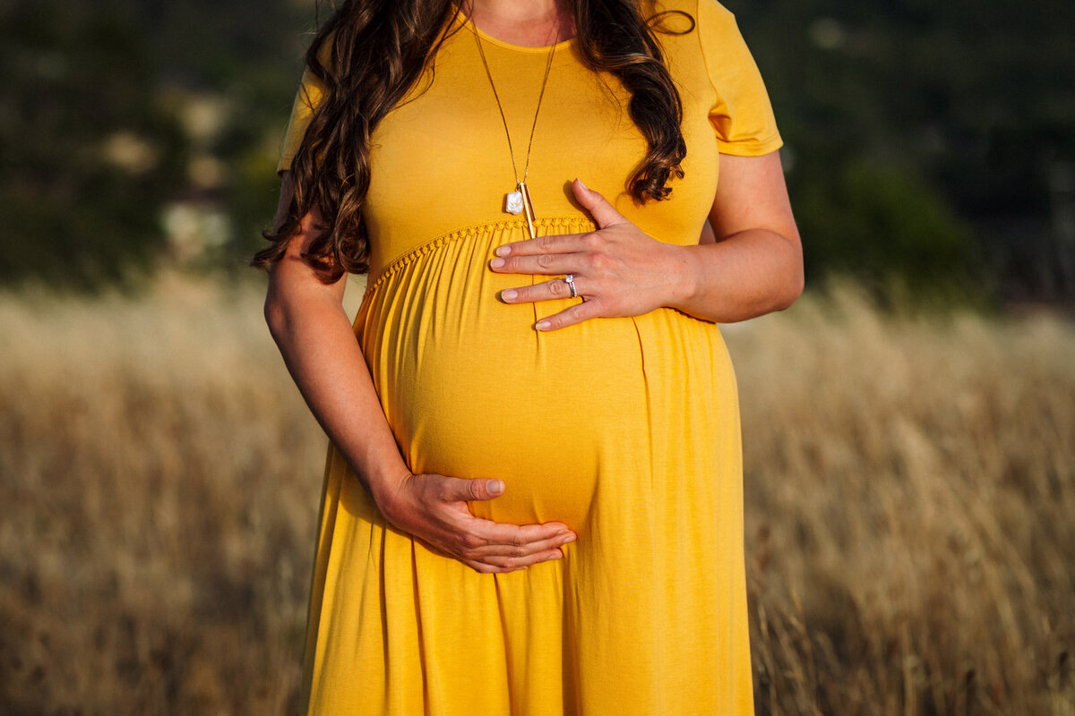 baby bump of pregnant woman in mustard color dress at golden hour standing in front of tall yellow grass