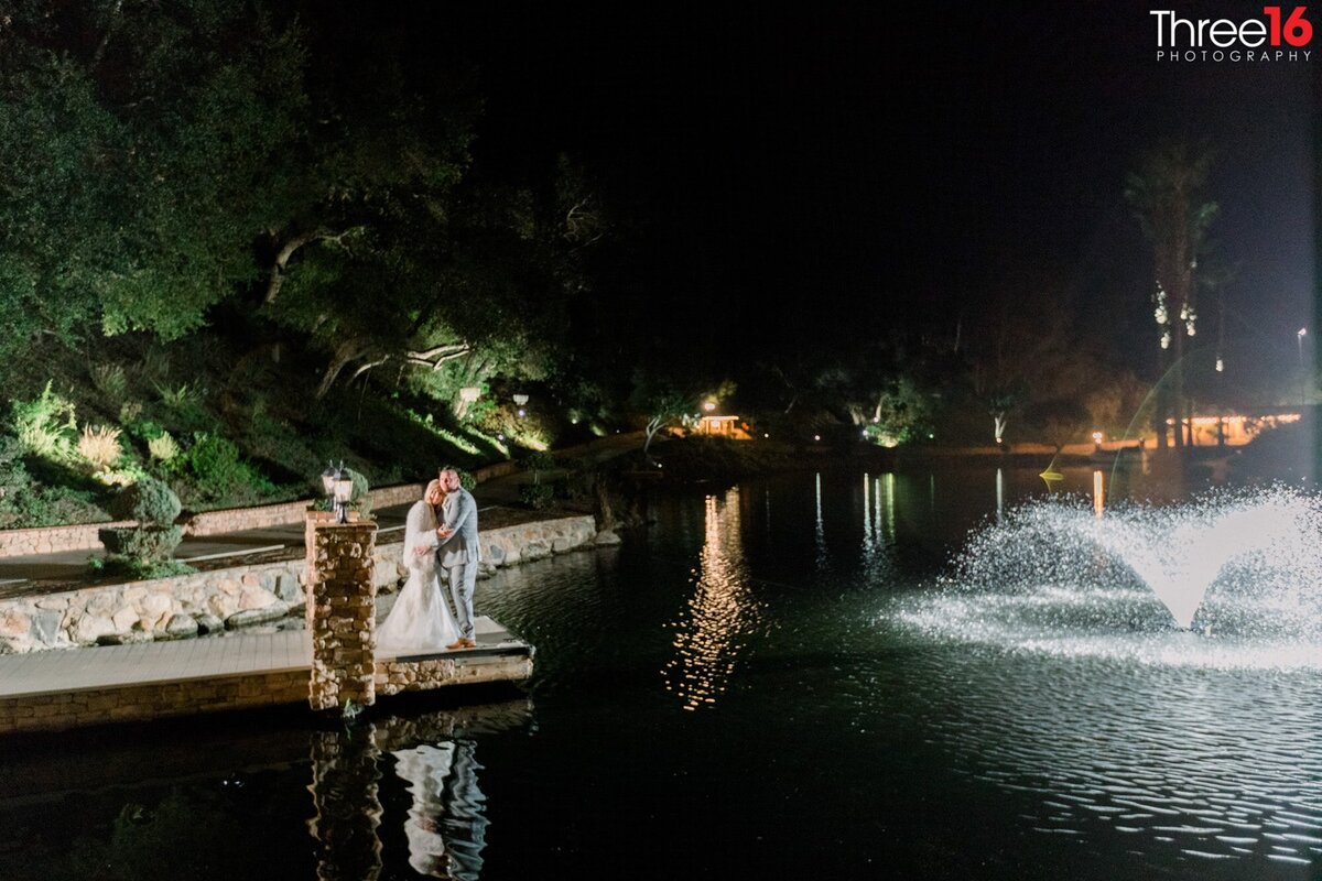 Night shot of Bride and Groom holding each other on a dock of the lake with water fountain shooting water into the air