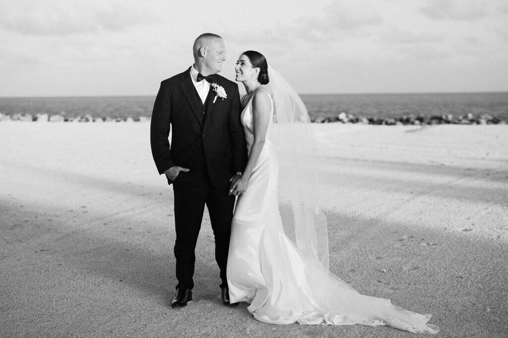 A heartfelt black-and-white image of the newlyweds smiling and embracing on the beach, radiating happiness on their wedding day. Captured by Claudia Amalia Photography.