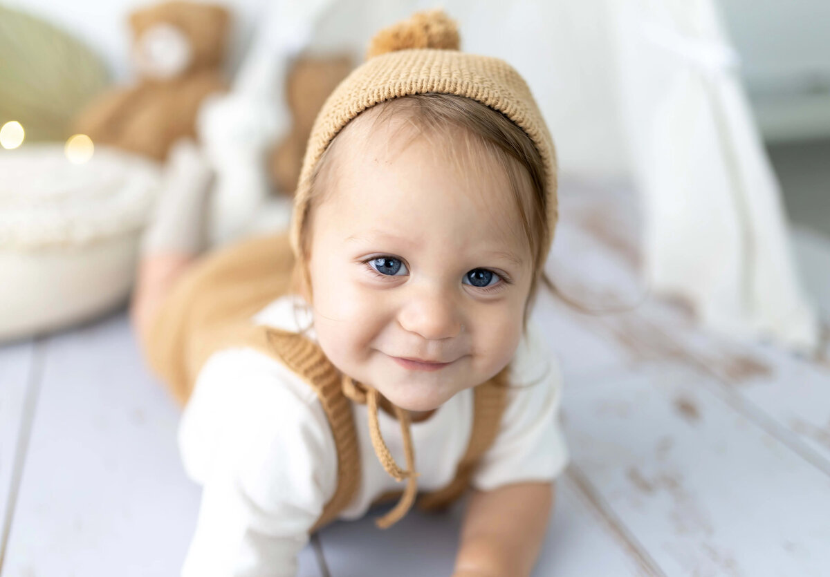 Smiling baby boy in brown bonnet crawling on whitewashed wood floor - milestone session with warm tones