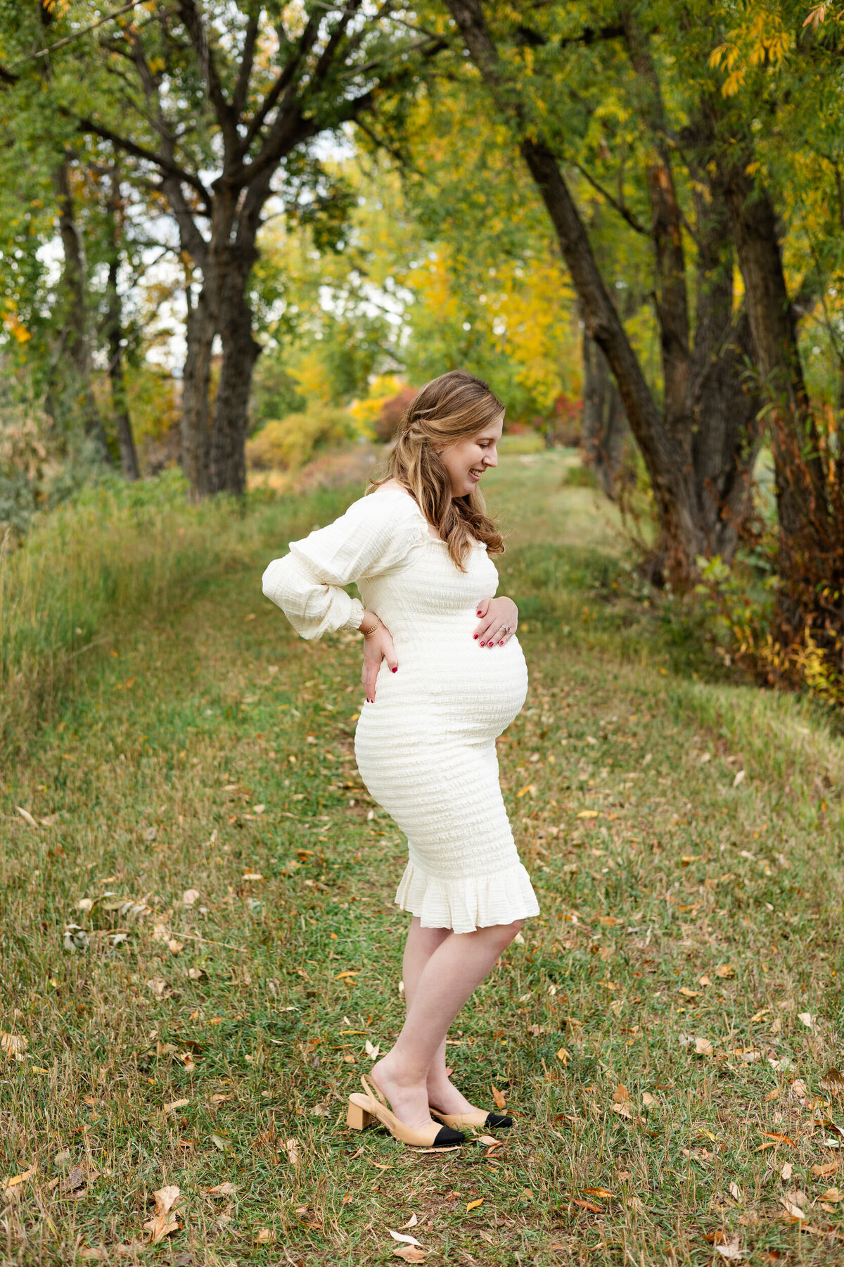 A pregnant woman stands on a wooded path with one hand on her belly and one hand on her back as she smiles down at her bump.