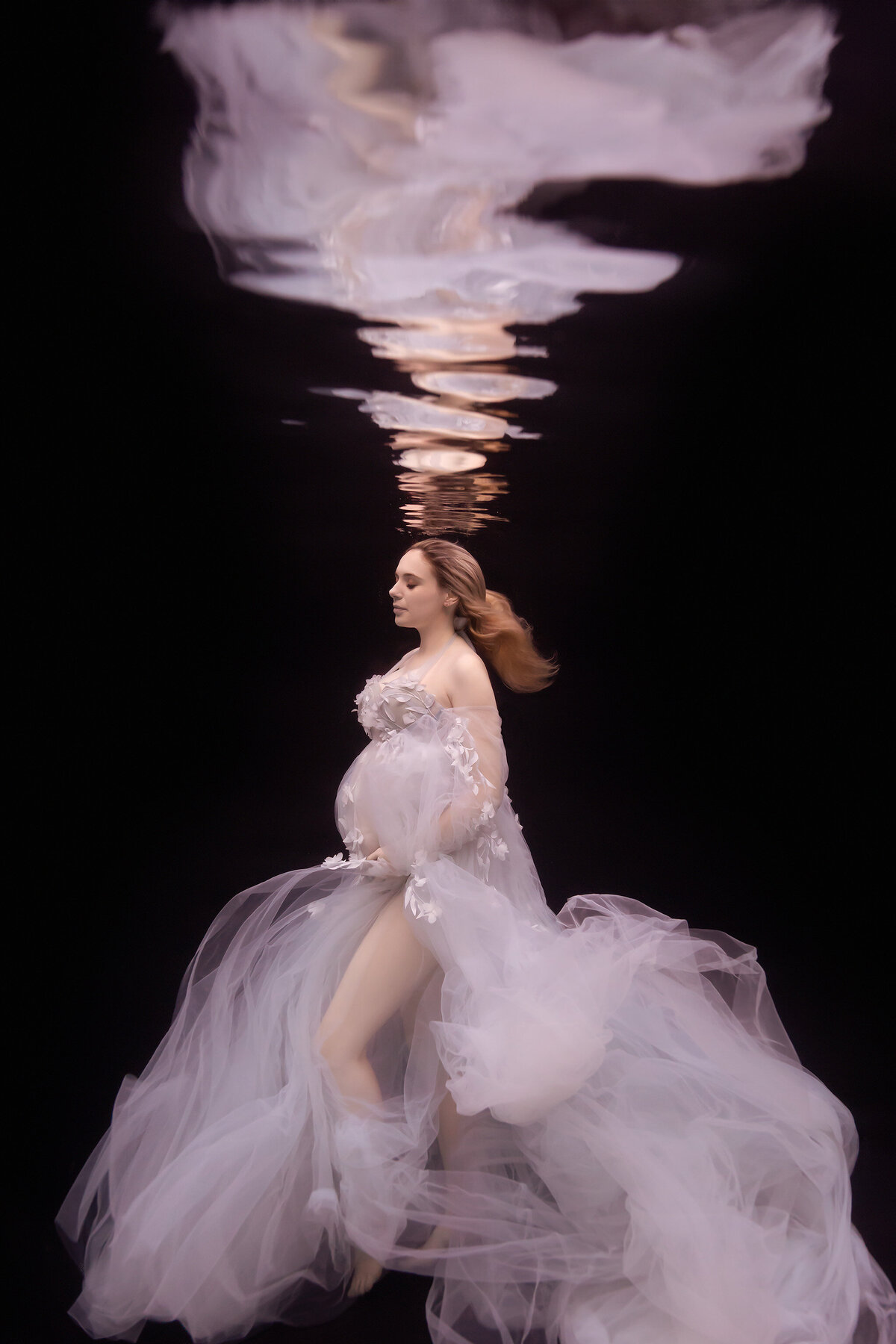 A captivating underwater maternity photo of a woman in a flowing white gown adorned with floral appliqués, her hair gently floating against a dark backdrop. The serene scene highlights her elegant silhouette and delicate reflection above, creating a timeless, ethereal feel. Dallas underwater photographer.