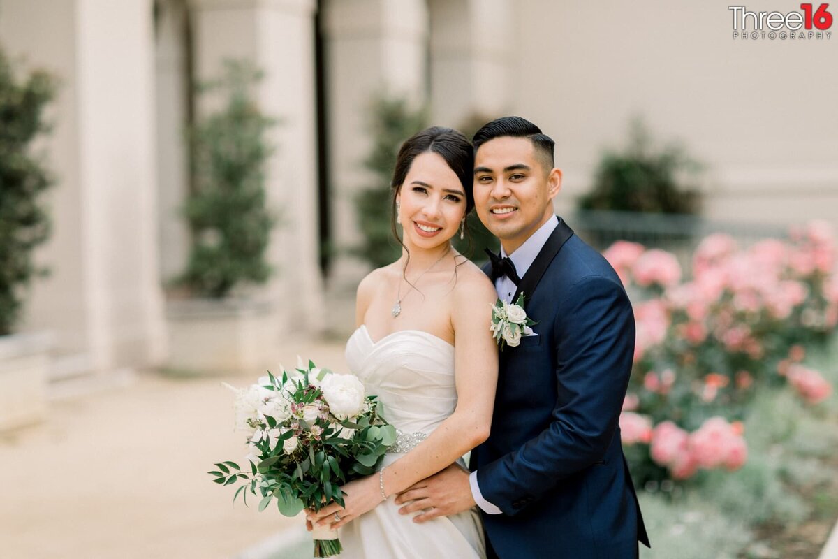 Groom holds his Bride from behind as they smile for the wedding photographer