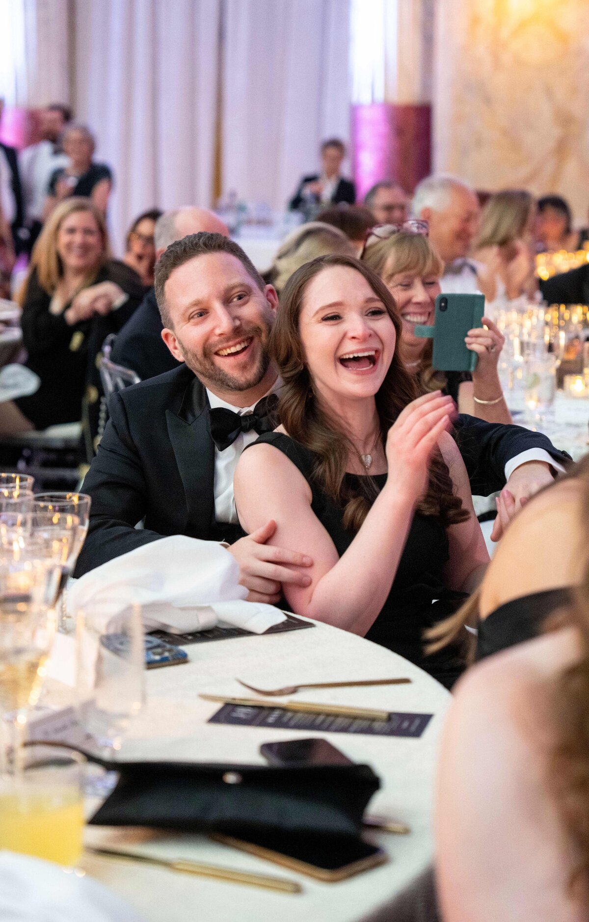 A joyful couple in formal attire sits at a banquet table, laughing amid a lively event. The man hugs the woman from behind. Guests in the background share the happy atmosphere, with some capturing moments on their phones.