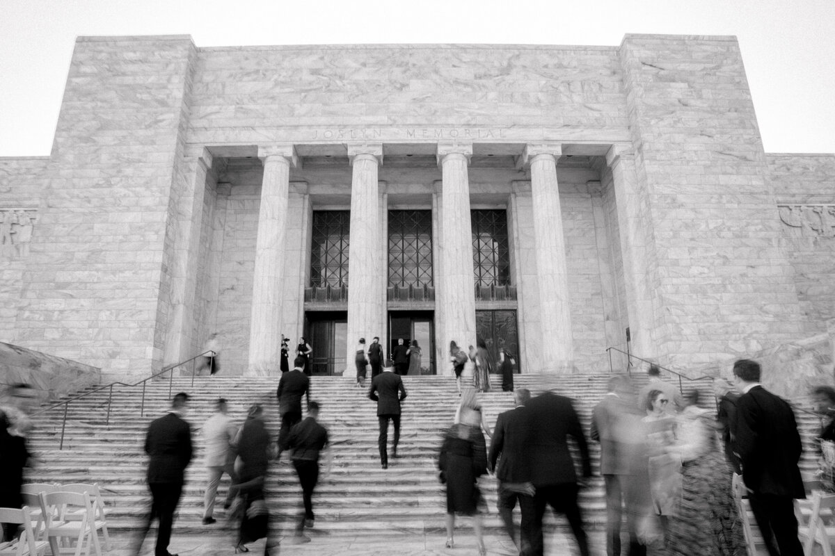 Black and white photo of a bride wearing a strapless dress with a bow and groom with a black tie tux walk hand in hand. Photo by Anna Brace, an Omaha wedding photographer.