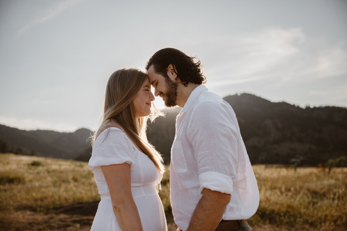 A couple in white outfits lovingly touch foreheads as the sun sets behind them at the Flatirons in Boulder, Colorado, surrounded by golden fields.