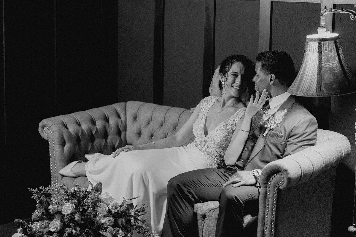 A candid black-and-white photo of the bride and groom on a tufted couch, smiling at each other.