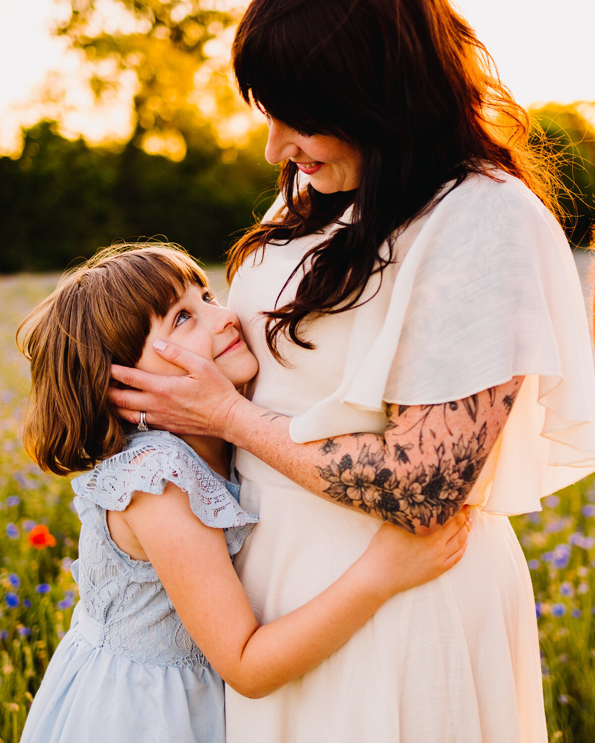 A girl in a light blue dress hugging a woman in a white dress with floral tattoos on her arm. The girl looks up and smiles, capturing a heartwarming moment of affection and joy.