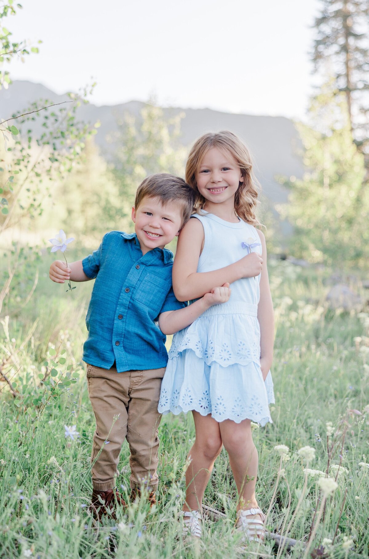 A brother and sister toddler pair lean on each other while smiling with wildflowers