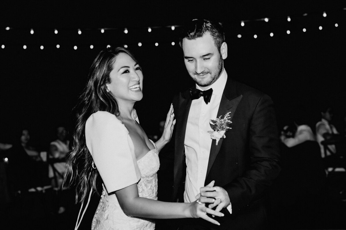 Bride and groom dance in black and white in front of string lights