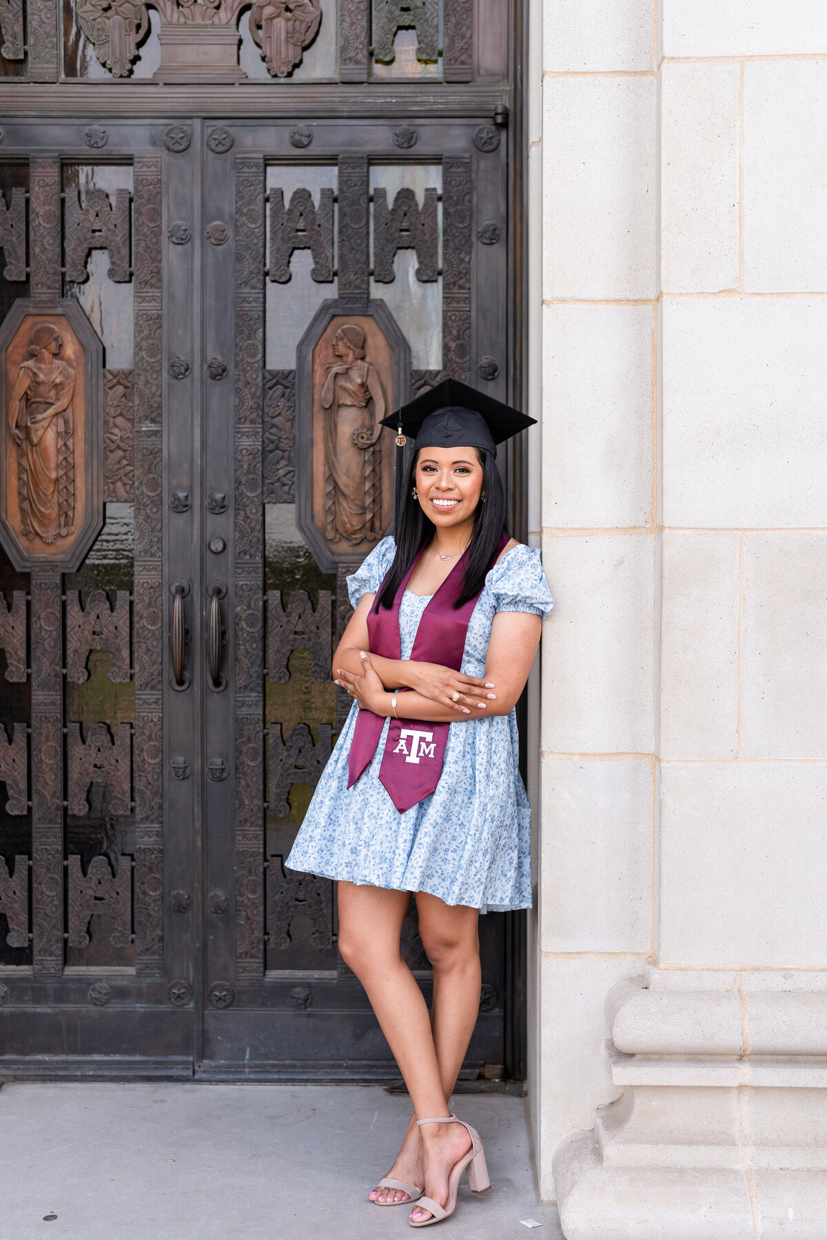 Texas A&M senior girl smiling and crossing arms while leaning against door of Administration building while wearing blue dress, maroon stole and grad cap