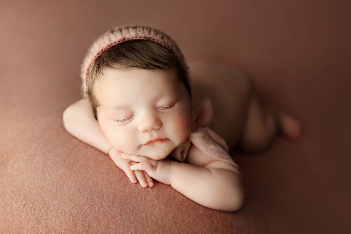 newborn laying on her tummy with her head on her hands with a pink bonnet on