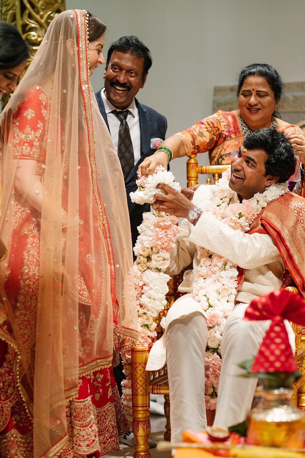 An Indian groom wearing a white sherwani with a red dupatta offers a white and blush floral varmala to a Hindu bride wearing a gold and red saree in Nashville, TN