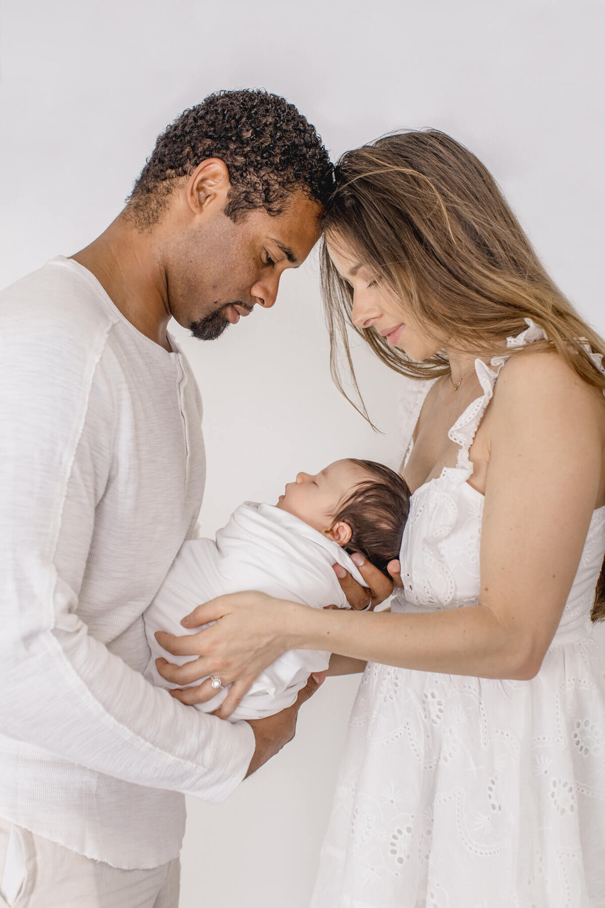 A couple lovingly gazes at their newborn baby, nestled in their arms. The father is wearing a white shirt, and the mother is in a white dress. The baby is swaddled in white, creating a serene and tender family moment.