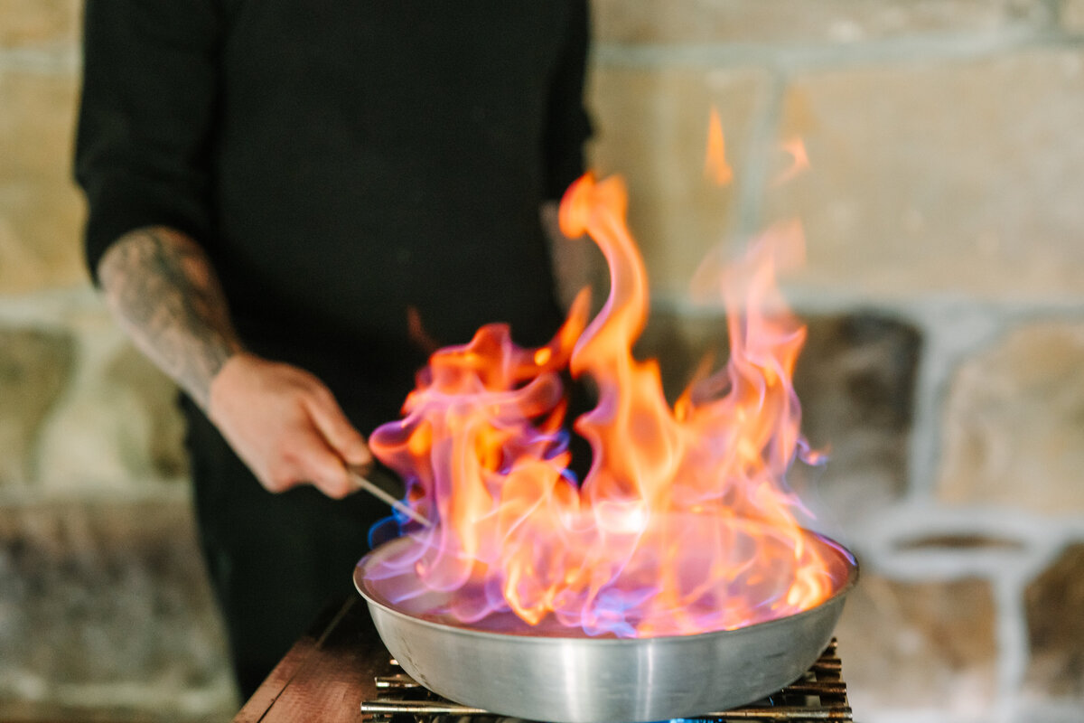 flambe flaming pan with a chef dressed in black in front of a stone wall at Willowbrook weddnig venue