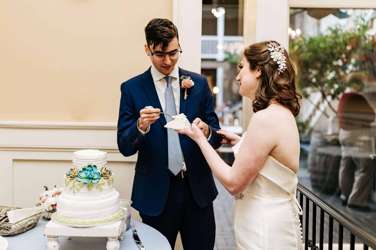 A bride and groom, dressed in wedding attire, smile as they cut and serve each other a piece of a two-tiered white wedding cake during an indoor event in a softly lit room.