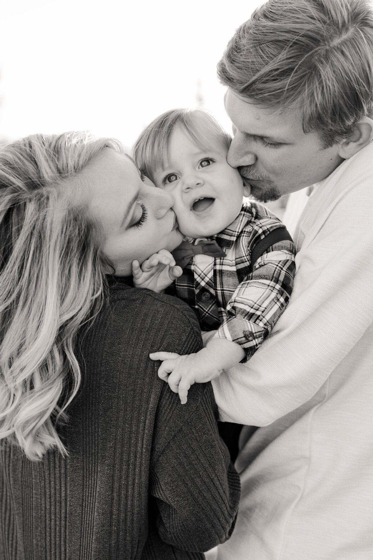 Black and white of Mom and Dad kissing son in between them with son smiling in awe