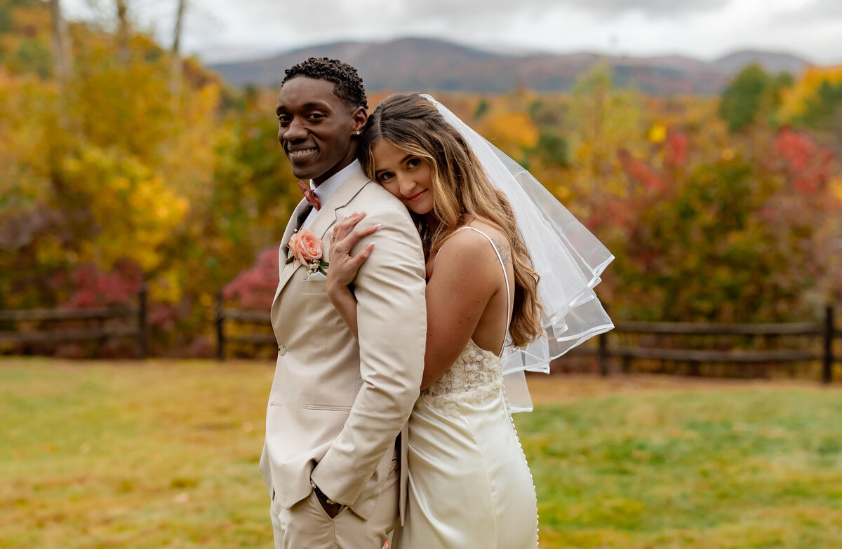 A bride and groom stand back-to-back amid the vibrant autumn foliage, captured beautifully by a New England Wedding Photographer. The groom, in a light suit with a peach boutonniere, and the smiling bride in her white gown, pose against a backdrop of colorful trees and mountains.