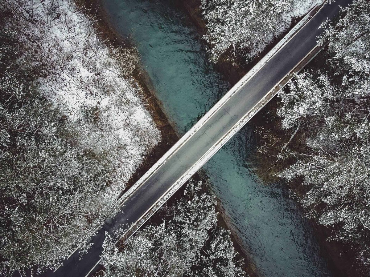 Minimalistische Winterlandschaft mit schneebedecktem Wald und klarer Brücke über türkisfarbenem Fluss – moderne Luftaufnahme von Atelier Schulte – professionelle Landschaftsfotografie in Kärnten.