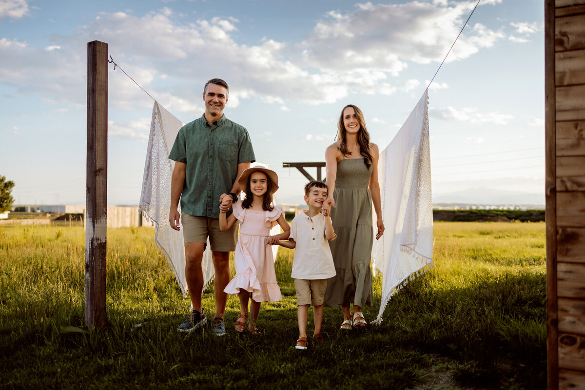 Family of four walk hand in hand in between two blankets hanging to dry on a family farm for their family photo session.