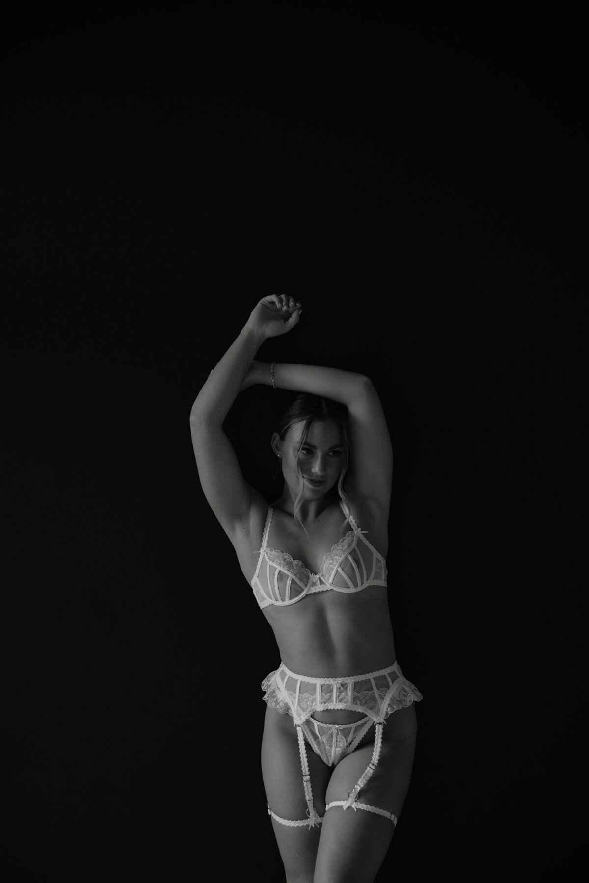 Black and white photo of woman in lingerie with her arms above her head standing in front of a black wall
