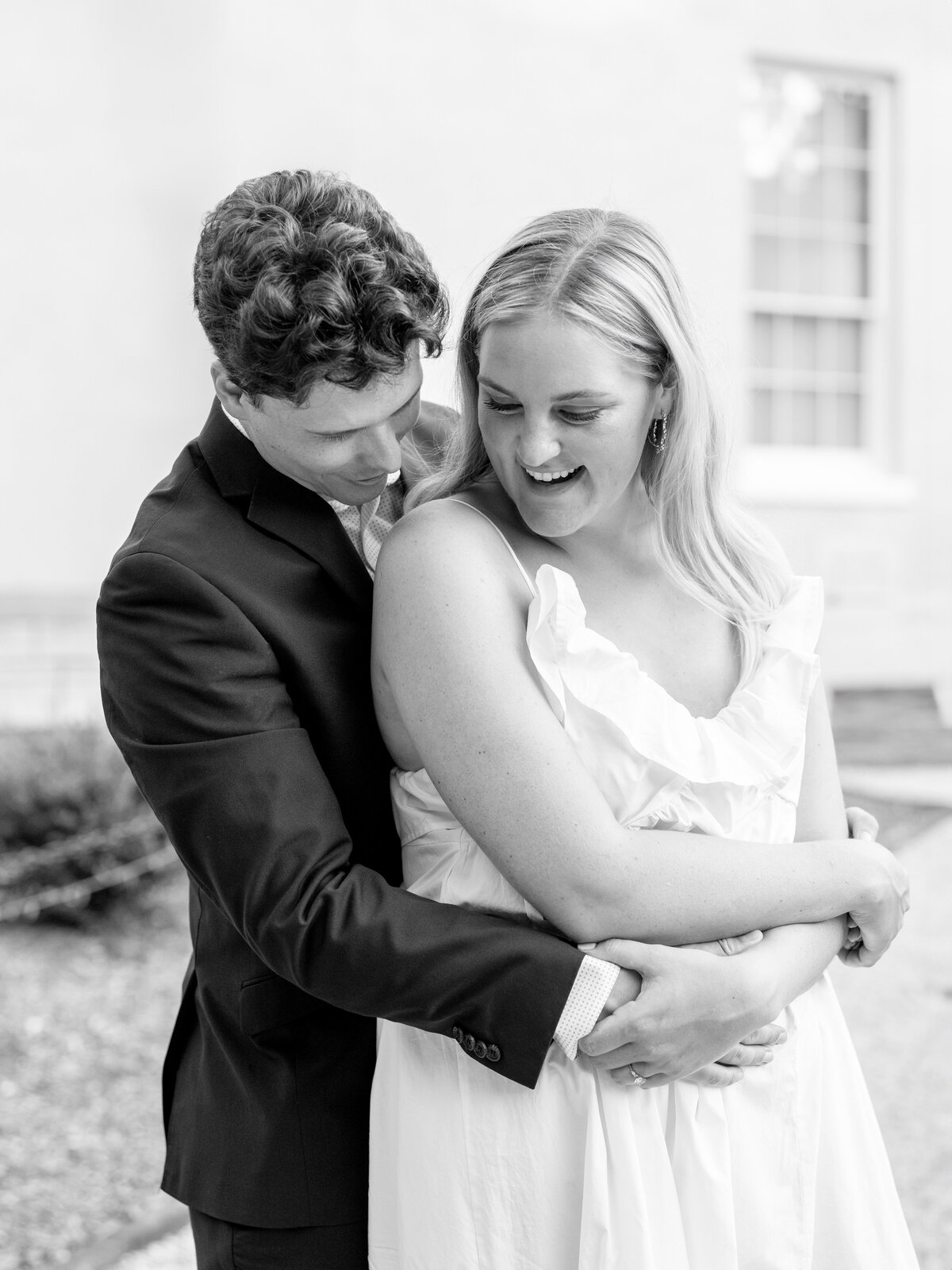 A black-and-white photo of a couple embracing. The man, wearing a suit, stands behind the woman, who is in a white dress. They are outdoors, against a light backdrop with a window visible. Both are smiling and appear happy.
