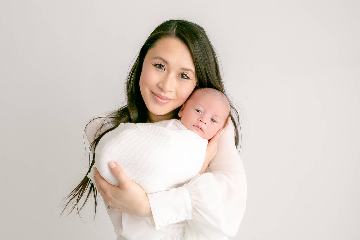 Mama with long black hair in loose curls. She is holding her baby up by her face. Her baby's eyes are open and he is wrapped in white. Mom has a soft smile on her face.