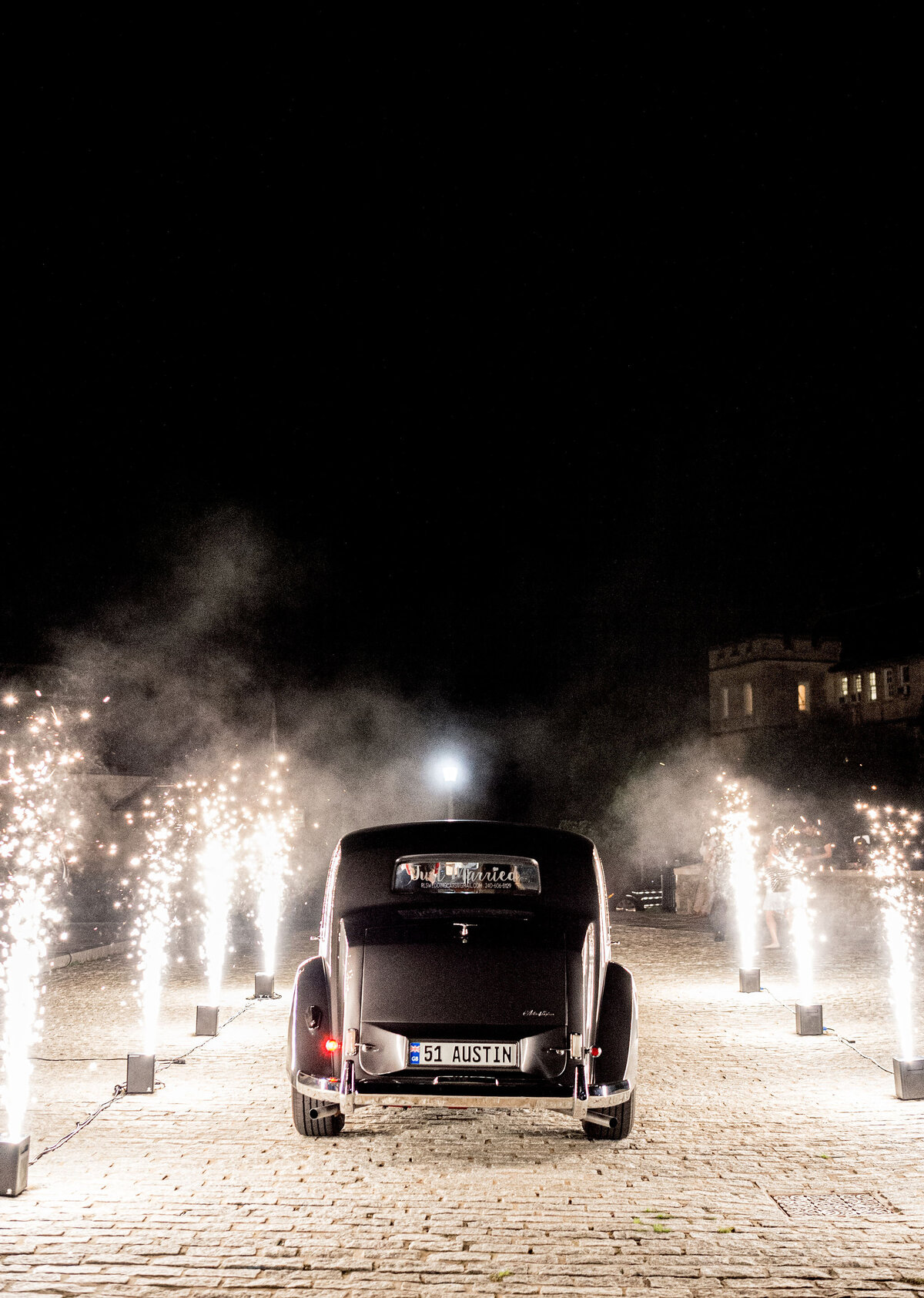 A classic car with the license plate 51 AUSTIN drives down a brick path at night, surrounded by sparklers on both sides. The scene is festive and illuminated by the bright light of the sparklers.