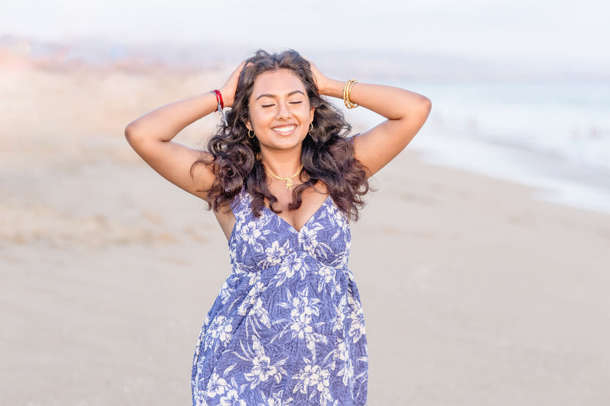 A woman in a blue floral dress stands on a beach, smiling with her eyes closed. She has her hands in her hair and the ocean is in the background under a clear sky.