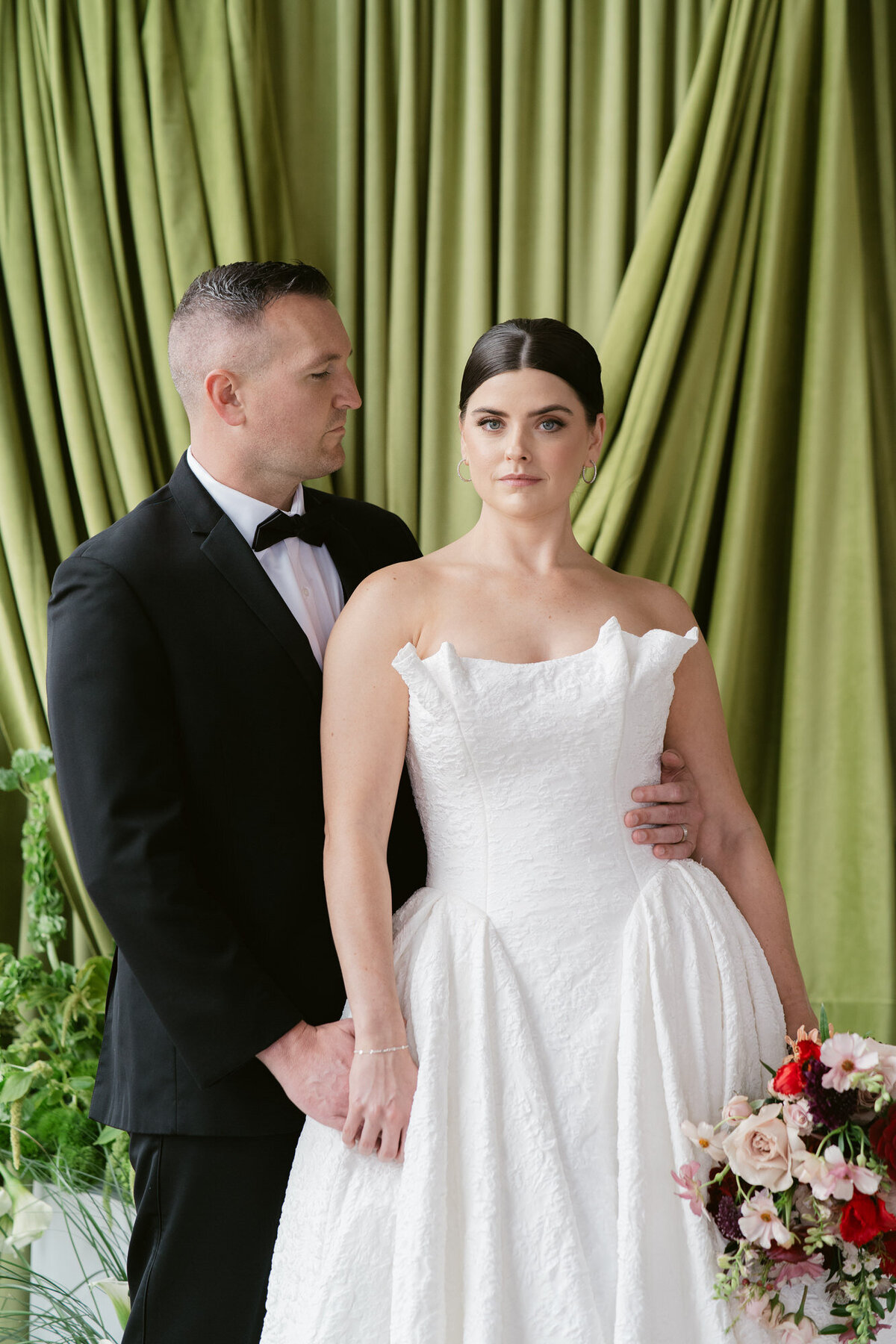 Groom looks at bride as she looks at the camera with an olive green drapery behind them.