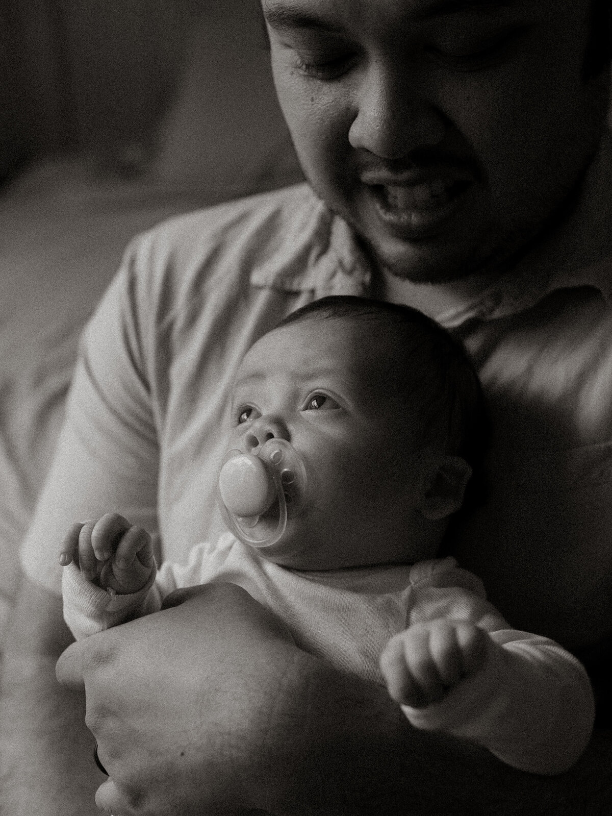 Black and white photo of a father lovingly cradling his newborn with a pacifier, both gazing upward in quiet connection.