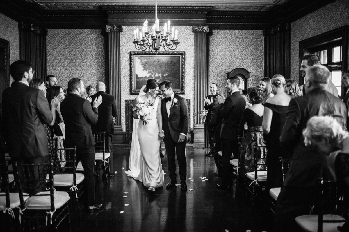 Black and white wedding ceremony photo with bride and groom walking down the aisle