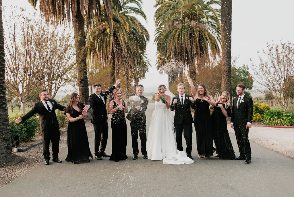 Bridal party celebrating with exploding champagne bottles, dressed in elegant black attire against a backdrop of palm trees.