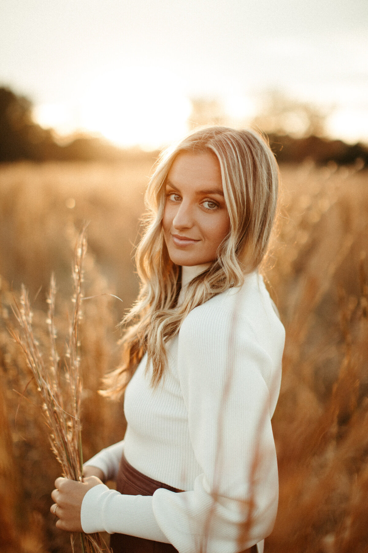 High school senior wearing cream sweater tucked into maroon leather skirt standing in a field on a fall day at golden hour