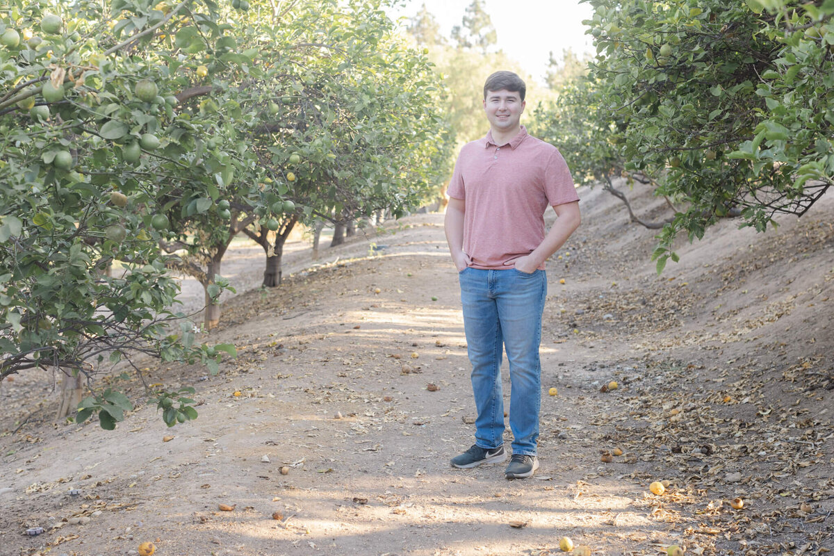 A young man wearing a salmon shirt and blue jeans standing with his hands in his pockets on a dirt path surrounded by lime trees.