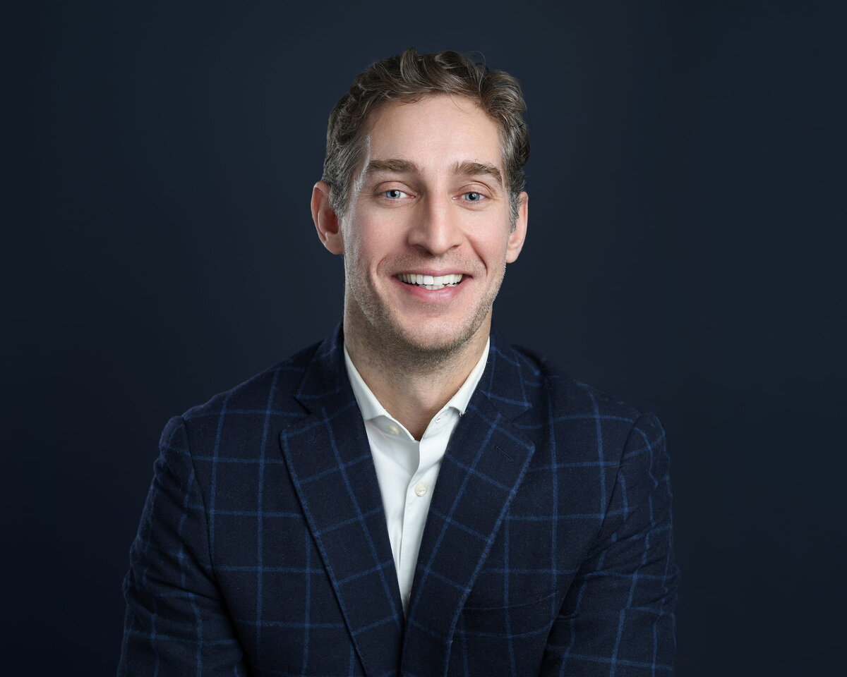 Professional headshot of an individual dressed in a stylish dark blue checkered blazer and crisp white shirt, posed against a rich dark blue backdrop. The subject exudes confidence and sophistication, making this image perfect for business branding and executive photography
