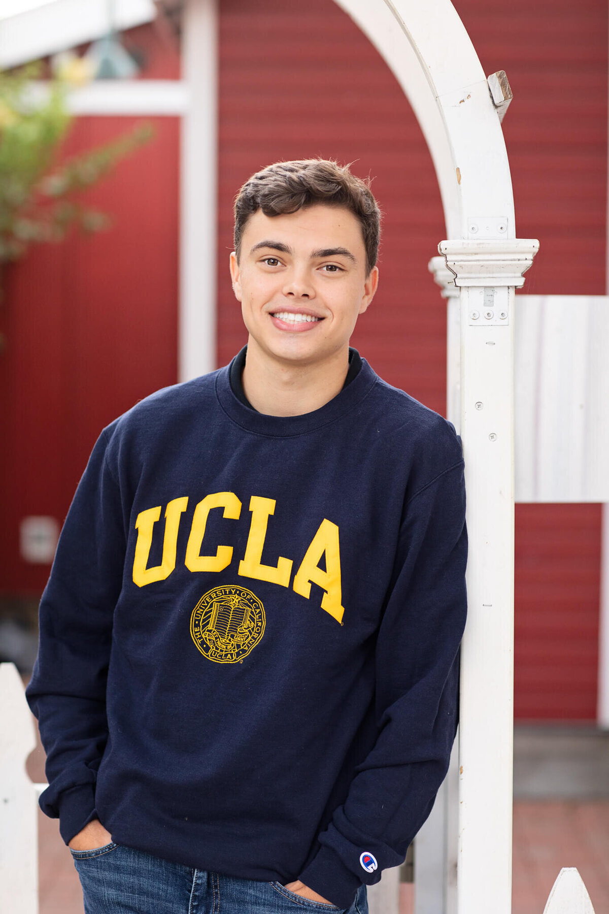 A person stands outdoors, smiling, wearing a navy blue UCLA sweatshirt. They are leaning against a white structure with a red background.