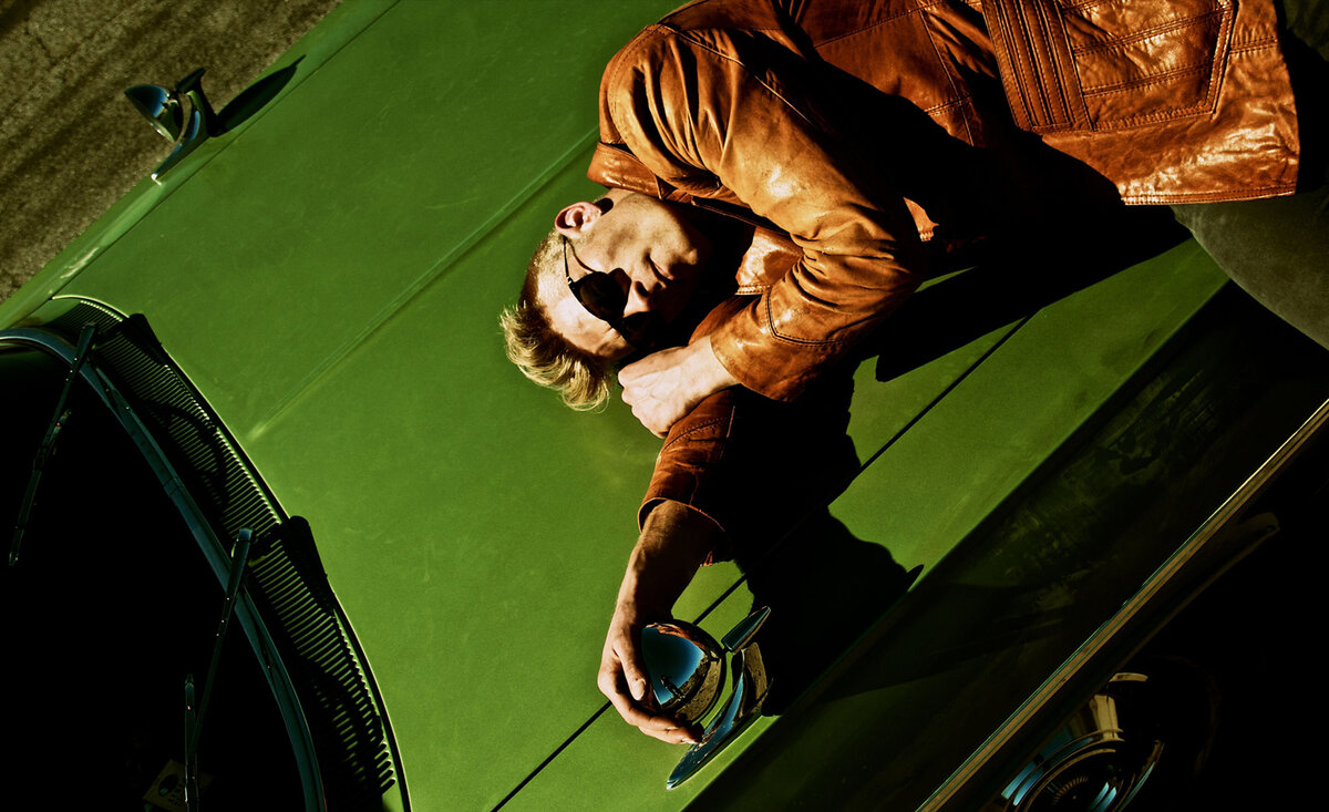 Male musician portrait Ryan Guldemond wearing tan leather coat lying against green vintage car hood