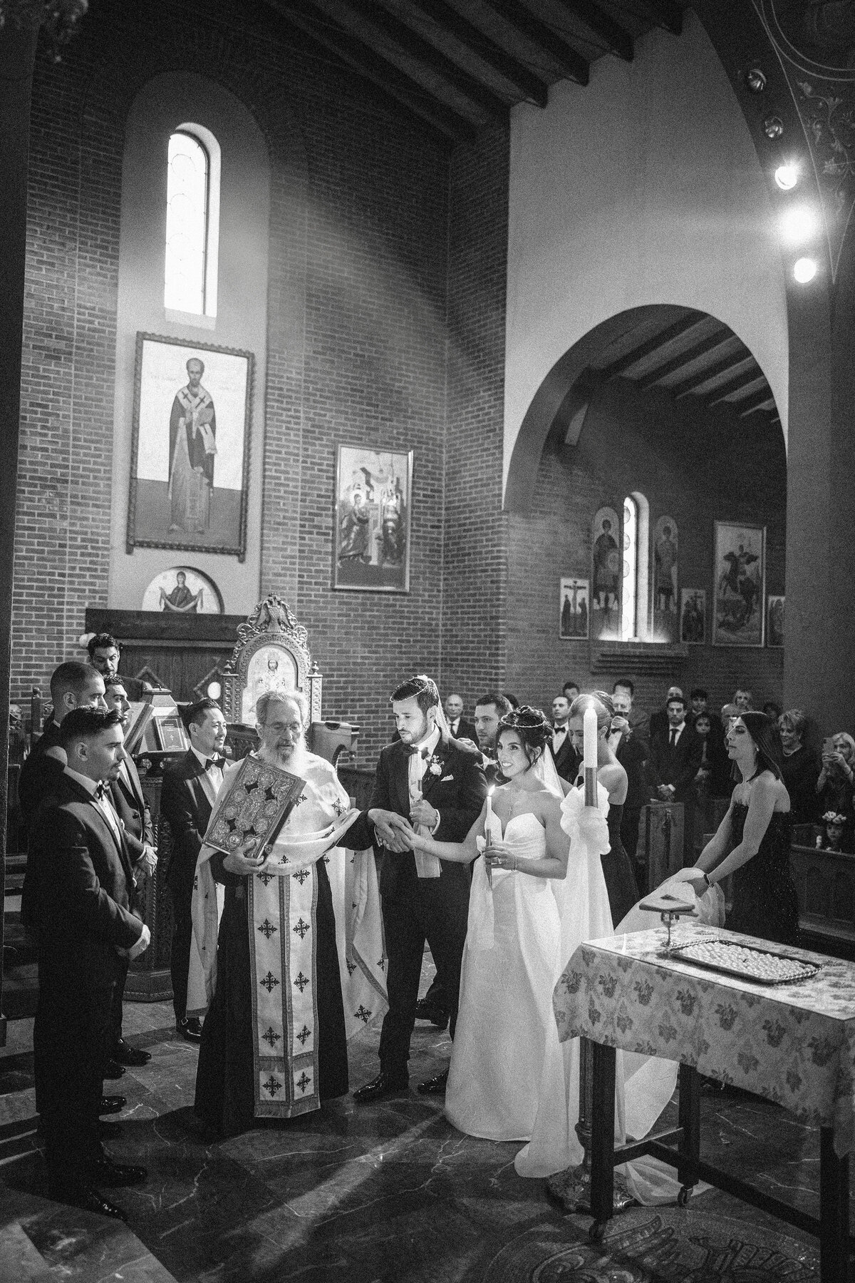 Black and white photo of a traditional Greek Orthodox wedding ceremony in progress, with the couple standing beside the priest holding candles and wearing ceremonial crowns, surrounded by family and friends inside St. Nicholas Church—capturing a sacred moment filled with ritual and reverence.