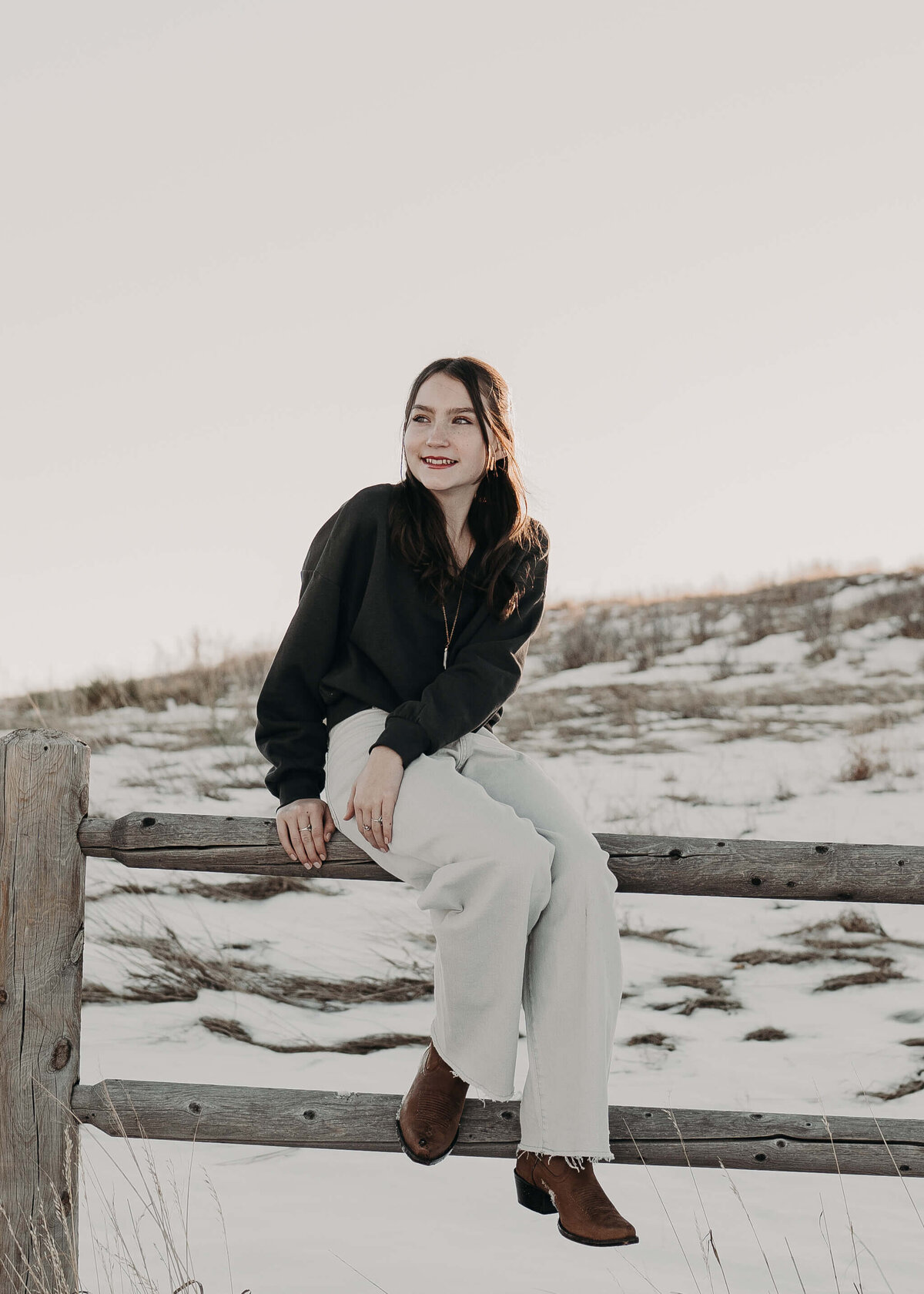 A beautiful preteen girl with fair skin and red lips sits on an old wooden fence while wearing cowgirl boots and cream colored pants, surrounded by snow and beautiful, tall grasses as the sunset paints a halo around her head.