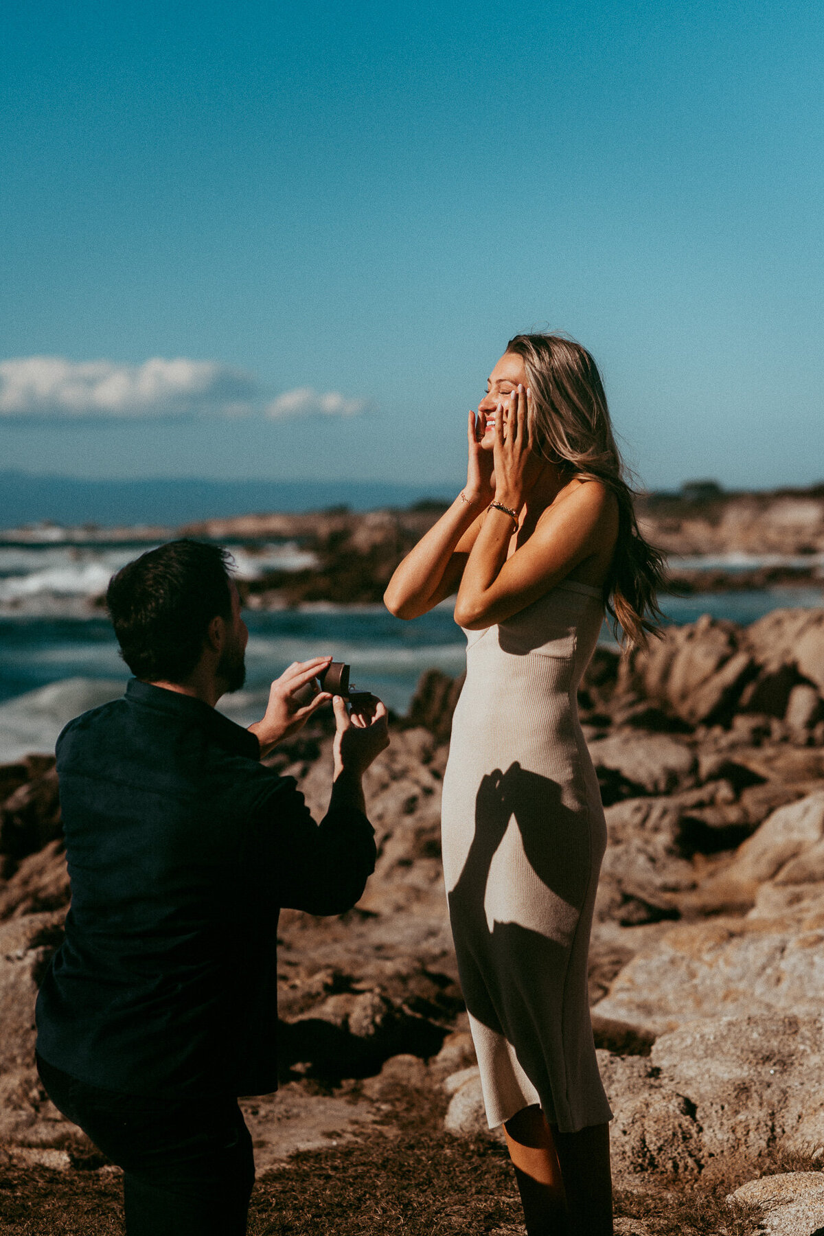 Man on one knee proposing on the rocks at Asilomar State Beach as she covers her face in surprise