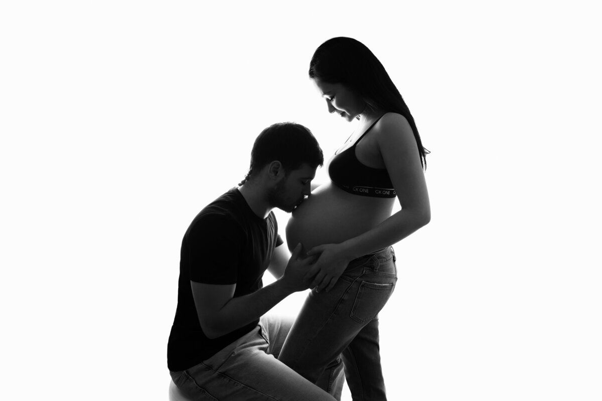 Husband sitting on a stool and kissing his pregnant wife's belly as she stands in front of him.