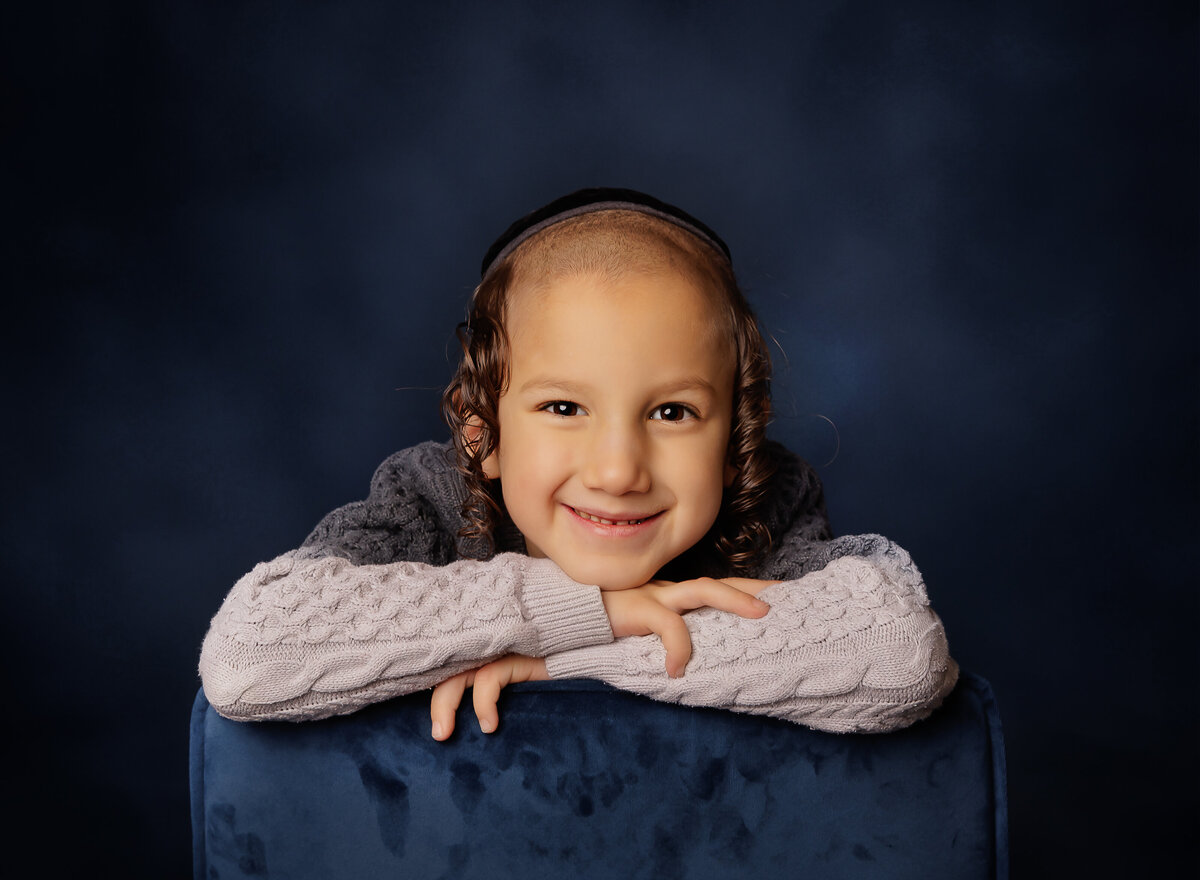 Little boy studio portrait is captured. He is sitting with his arms folded on the back of the chair and resting is chin atop of his hands. He is smiling at the camera. Captured by Brooklyn family photographer Chaya Bornstein